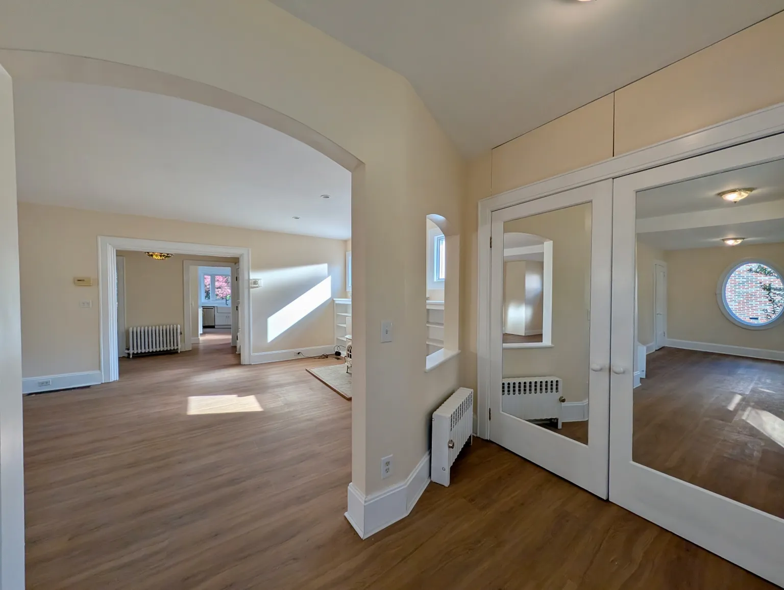 The entry looking through an archway and a line of rooms to a bright window at the back of the house, with mirrored French doors on the right reflecting the oval window