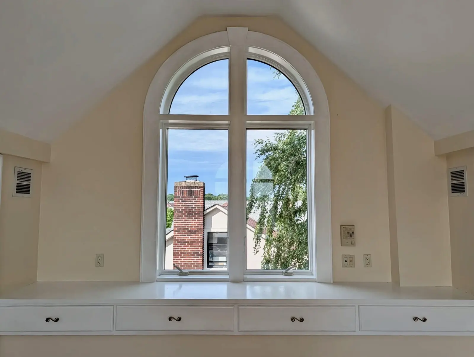 East-facing cathedral-arched window in the suite frames the sky under the vaulted ceiling, with a brass-handled built-in bank of drawers beneath the sill